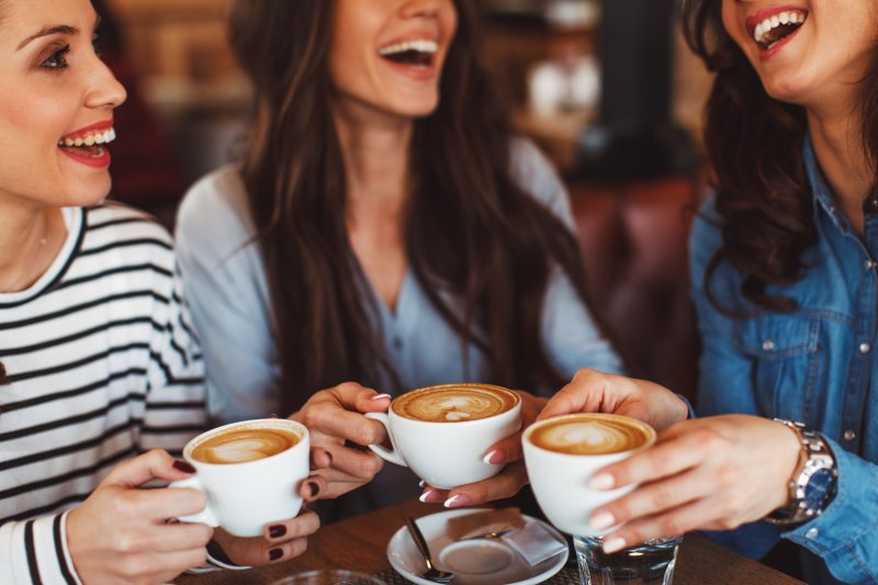 3 young women around a small wooden table holding hot lattes in white cups while smiling and laughing